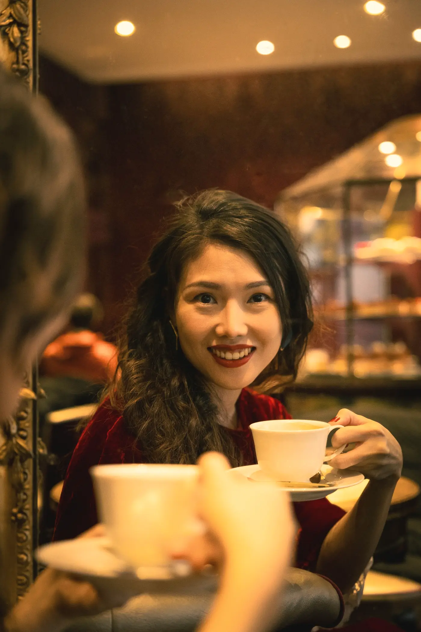A woman laughing across a café table, raising her cup to the person opposite